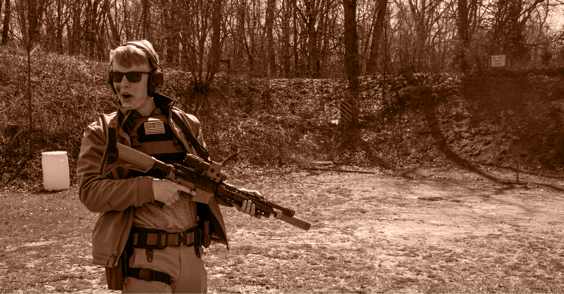 Person in tactical gear holding a rifle with EOTECH HWS sight and magnifier in an outdoor setting with trees and a wall.