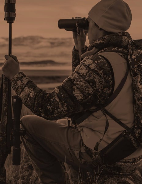 Person in camouflage gear using binoculars and a rifle with a EOTECH Vudu, sitting on the ground with a copper overlay