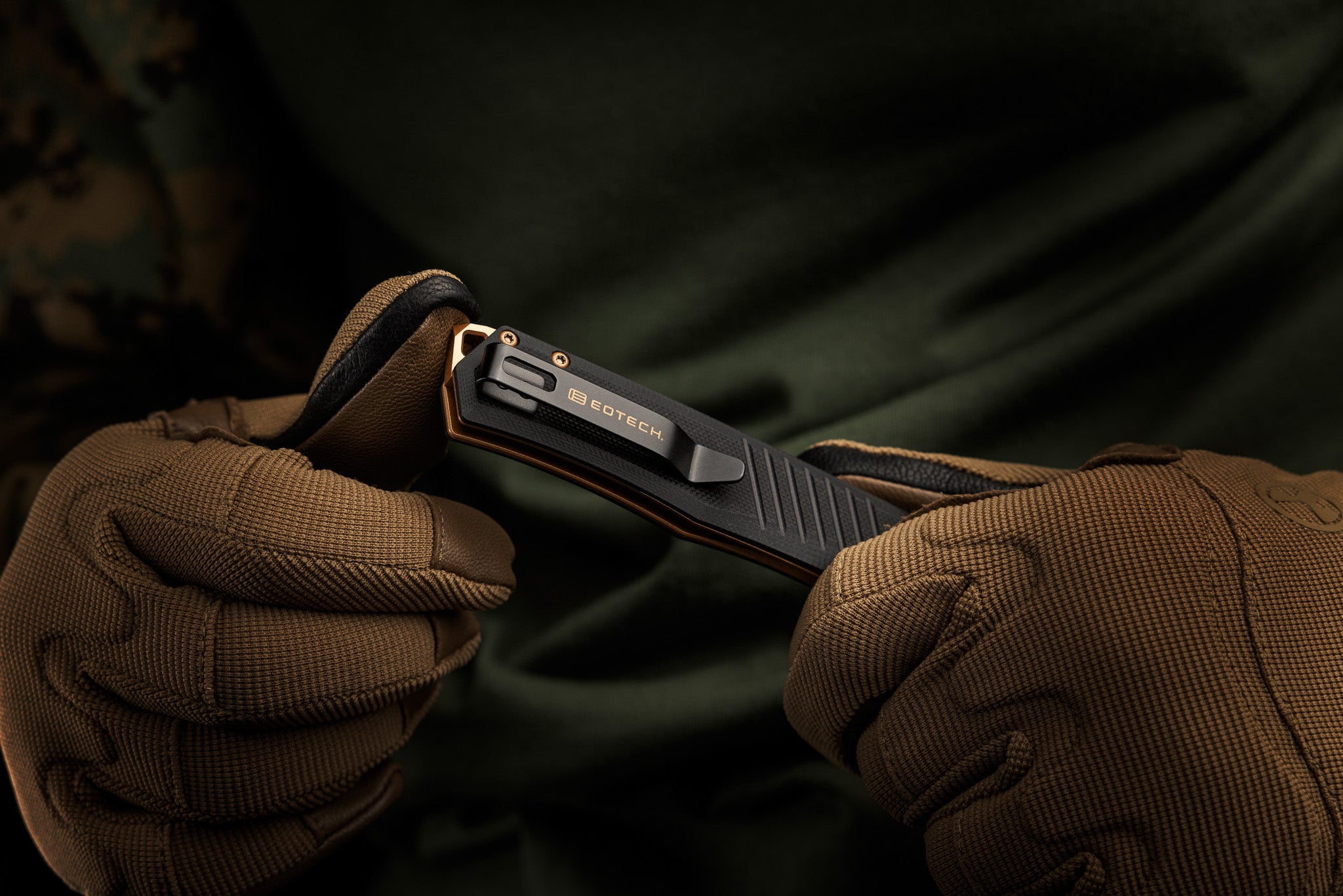 Person wearing brown gloves holding a black EOTECH folding knife with a dark background