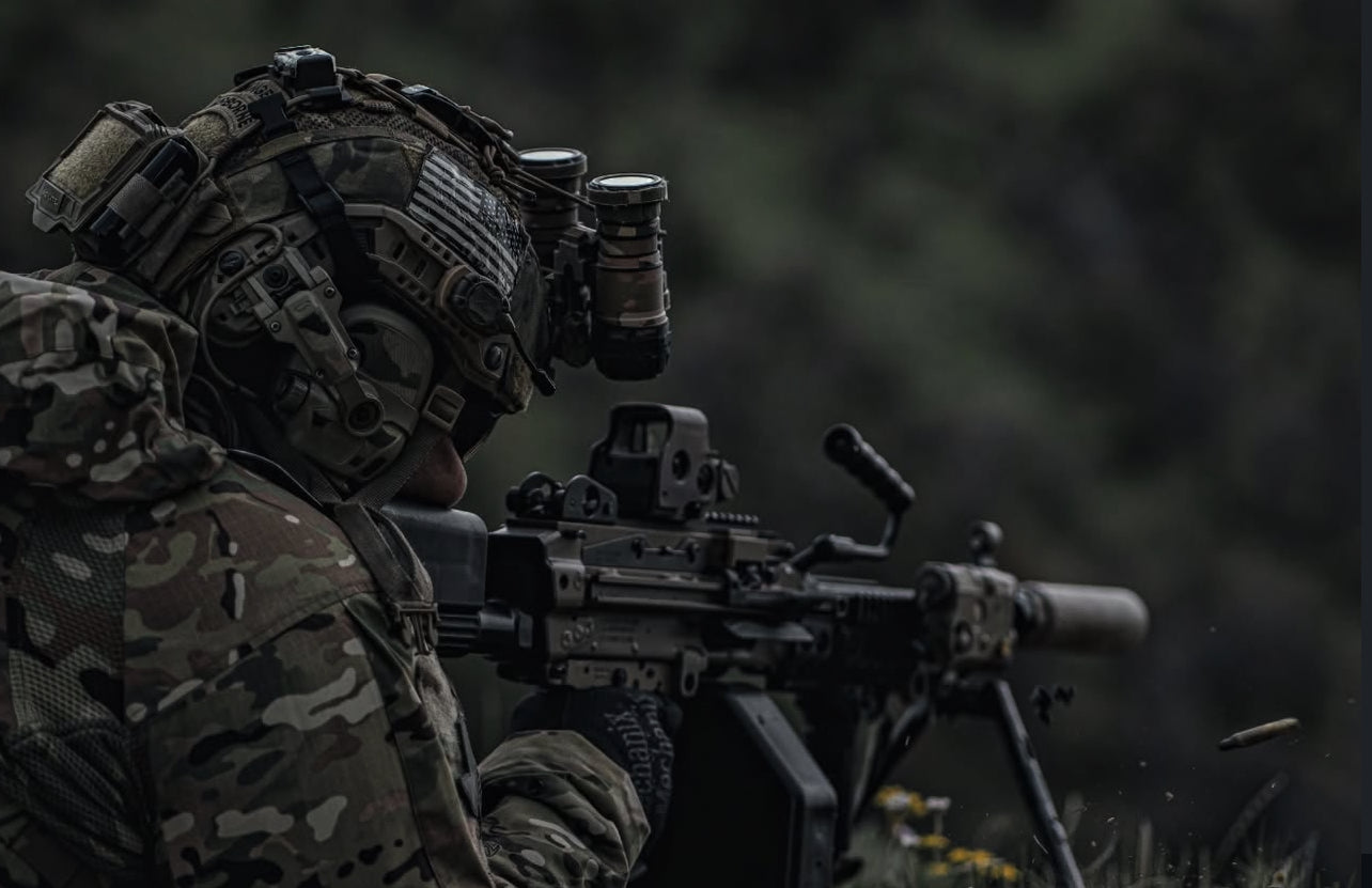 Soldier in camouflage gear with a rifle with EOTECH HWS and binoculars in a dark, forested area