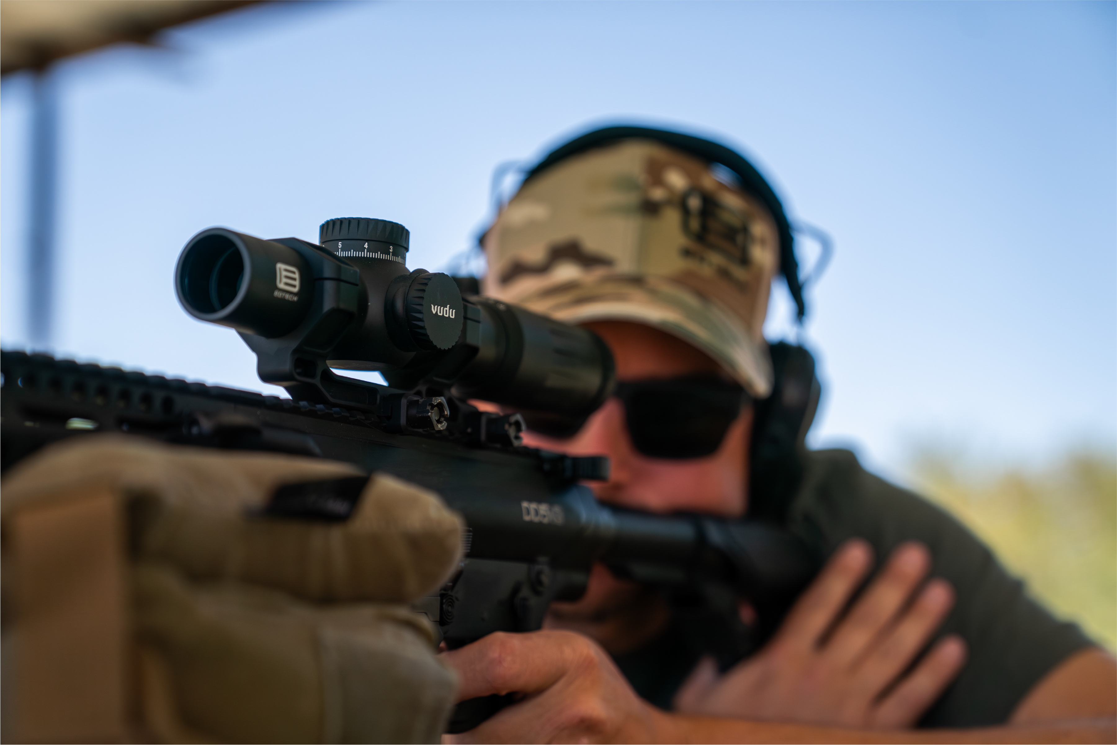 Person aiming a rifle with a EOTECH Vudu scope, wearing a camouflage hat and sunglasses.