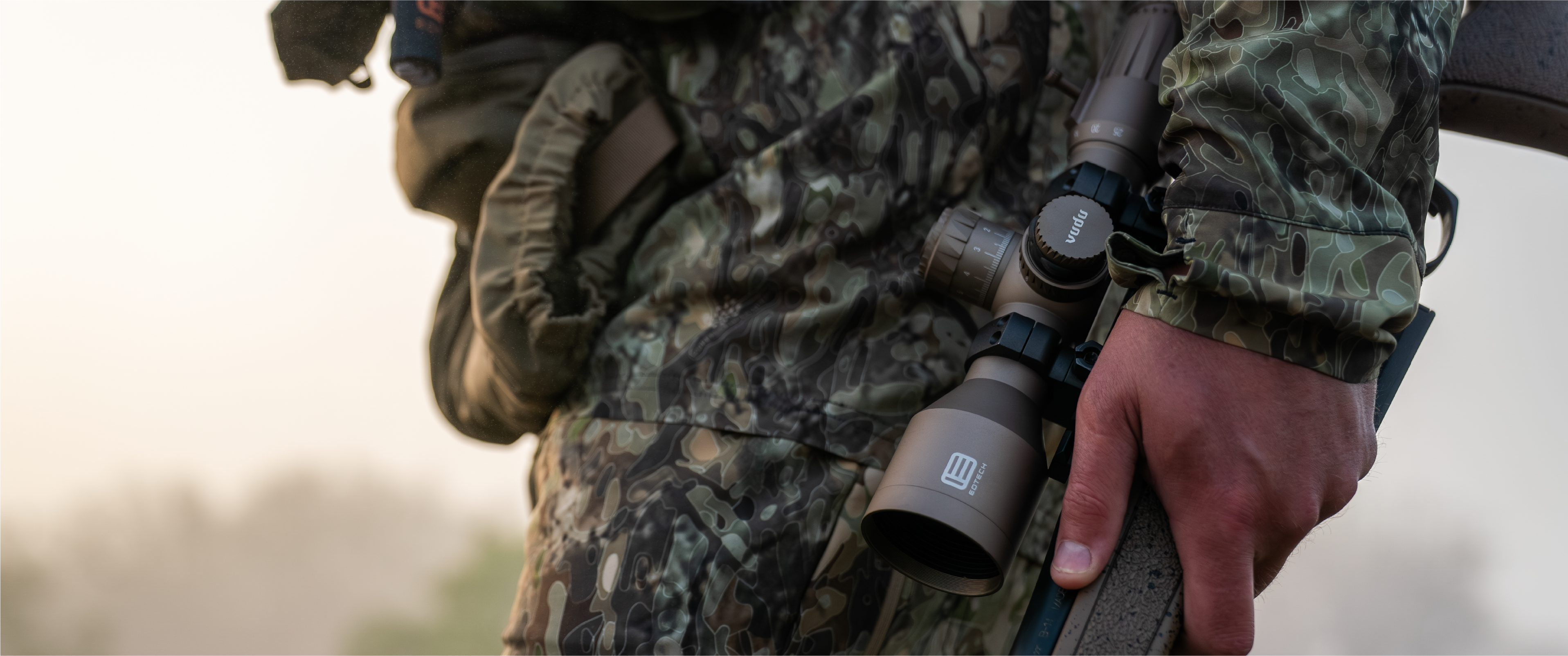 Person in camouflage holding a rifle with a tan EOTECH Vudu scope, outdoors.